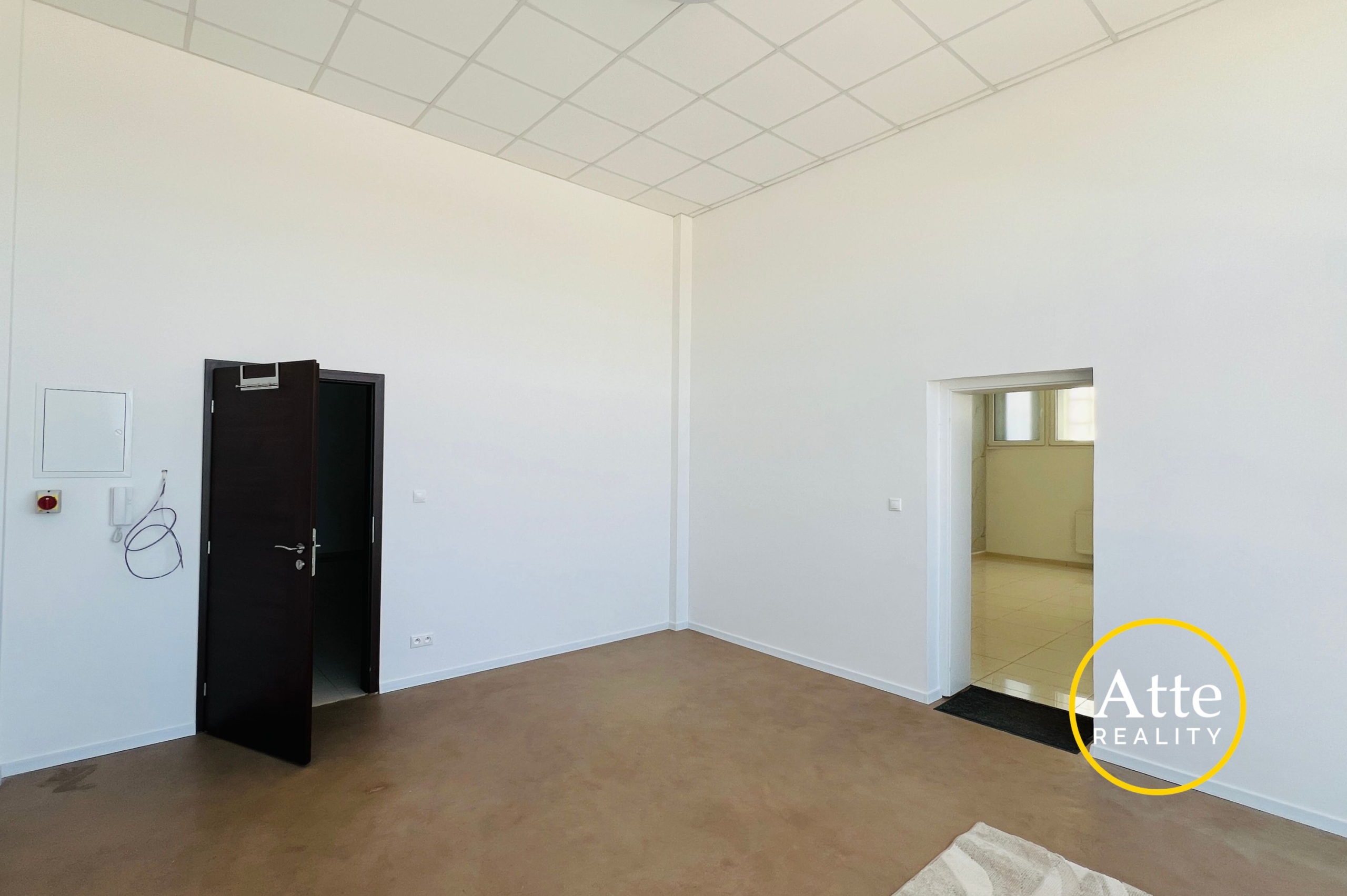 Empty office-like room with white walls, brown floor, a dark open door on the left, and a doorway to another room on the right.