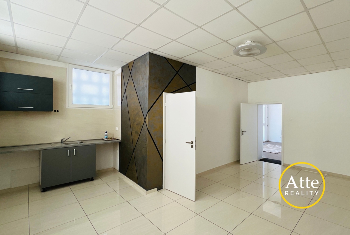 Empty modern kitchen or showroom with gray cabinets and stainless sink on left, geometric dark accent wall in center, white tiled floor and ceiling; Realty watermark in bottom-right corner.