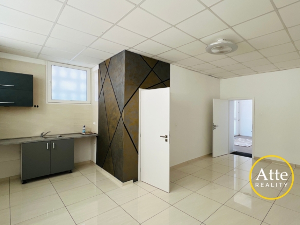 Empty modern kitchen or showroom with gray cabinets and stainless sink on left, geometric dark accent wall in center, white tiled floor and ceiling; Realty watermark in bottom-right corner.
