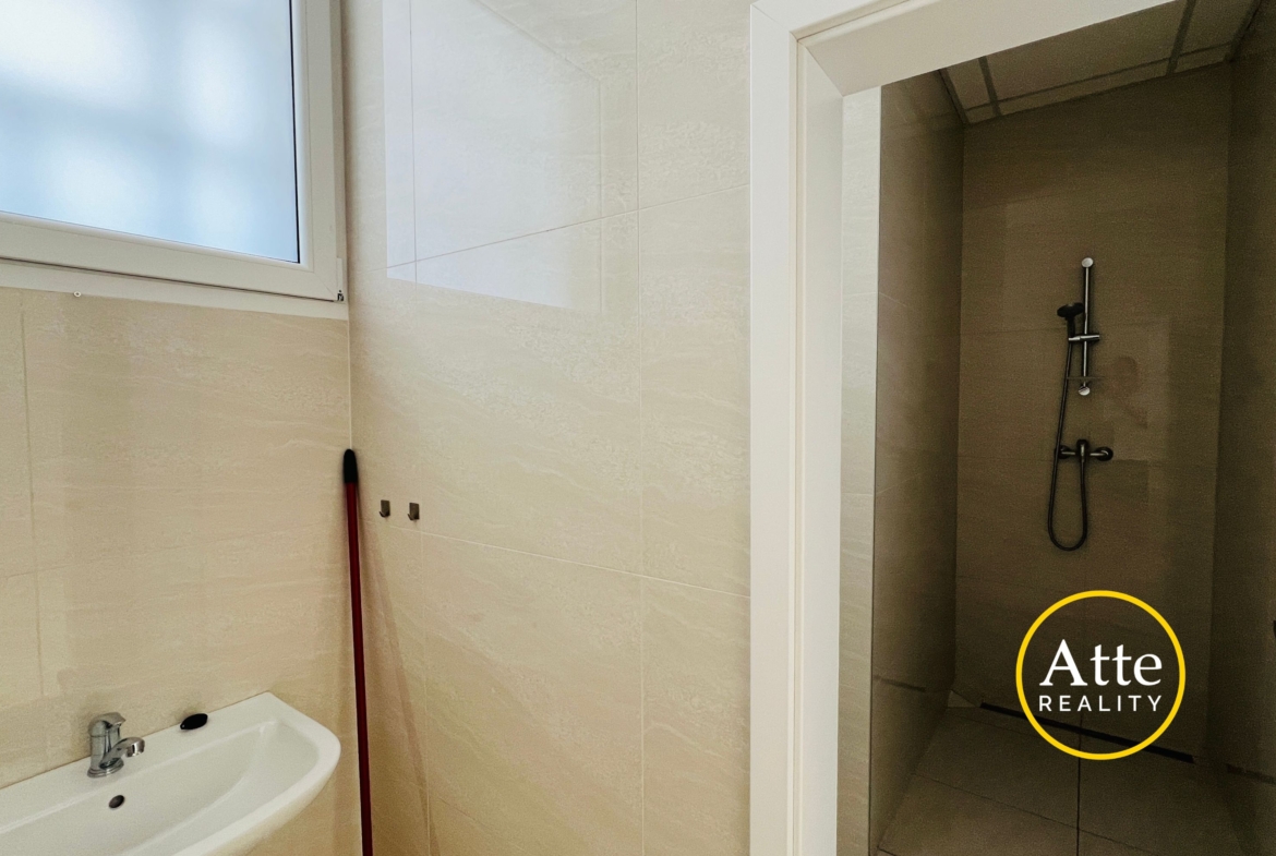 Bathroom with beige tiled walls, a white sink in the foreground, a frosted window to the left, a red mop leaning by the wall, and a glass shower enclosure on the right side.