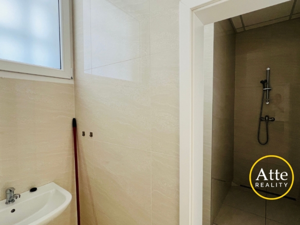 Bathroom with beige tiled walls, a white sink in the foreground, a frosted window to the left, a red mop leaning by the wall, and a glass shower enclosure on the right side.