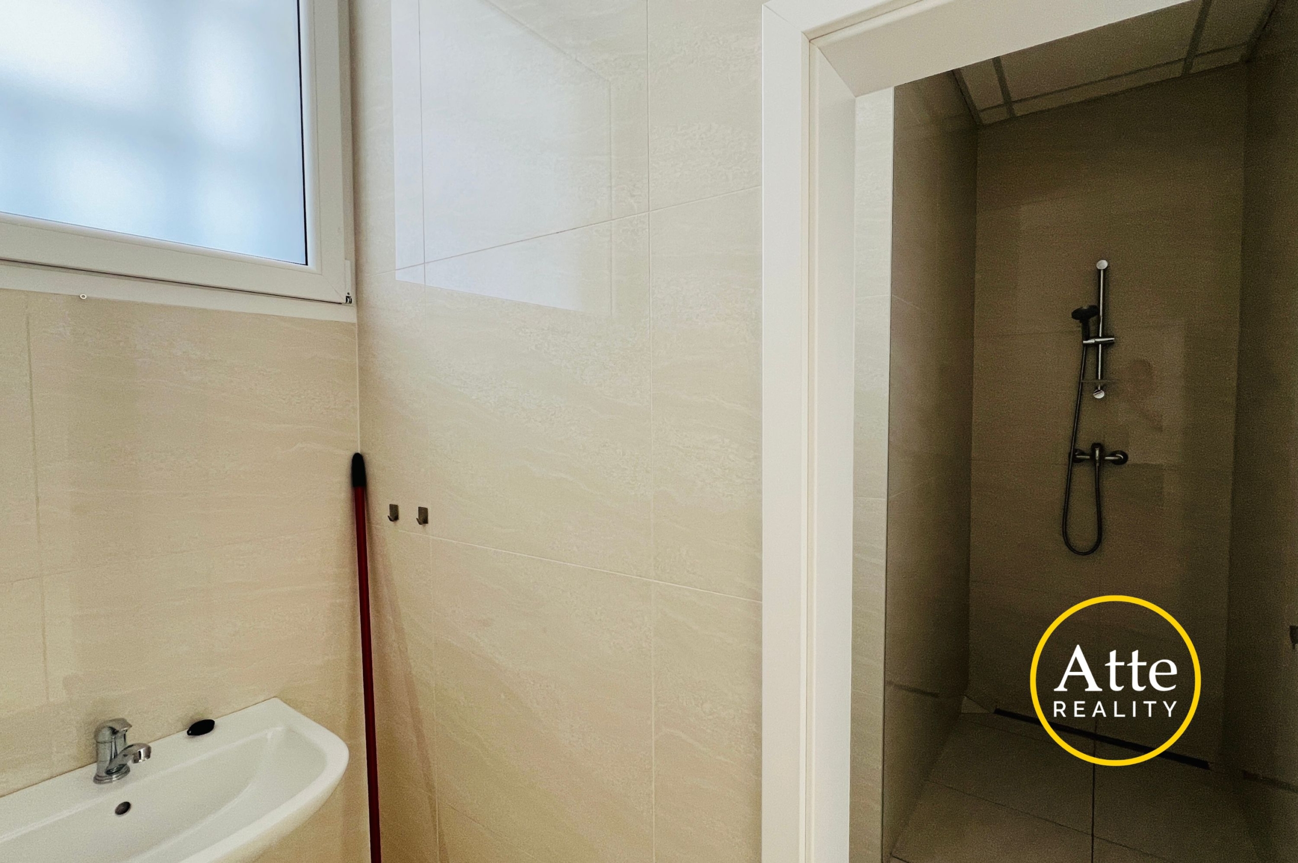 Bathroom with beige tiled walls, a white sink in the foreground, a frosted window to the left, a red mop leaning by the wall, and a glass shower enclosure on the right side.