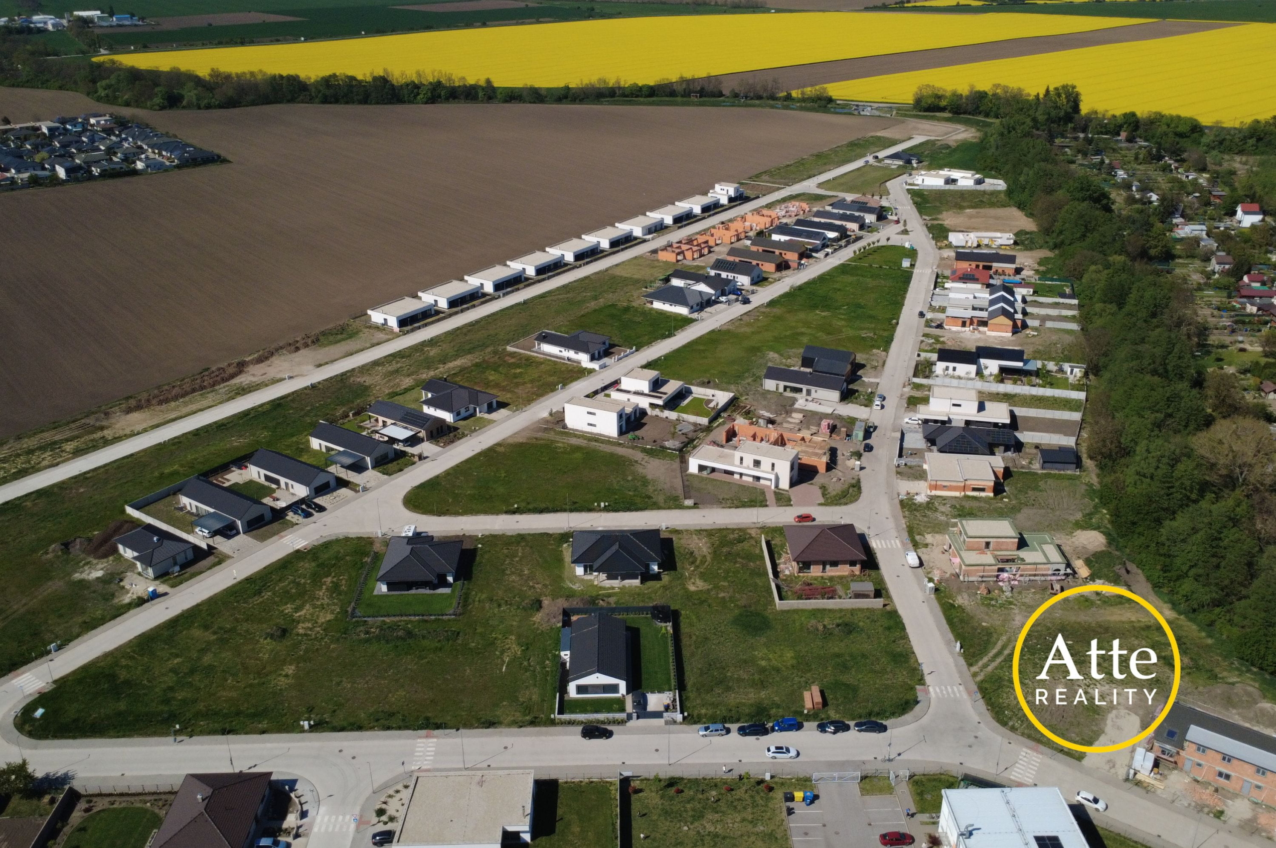 Aerial view of a new suburban neighborhood with rows of houses along streets, brown fields nearby and a bright yellow crop field in the distance.
