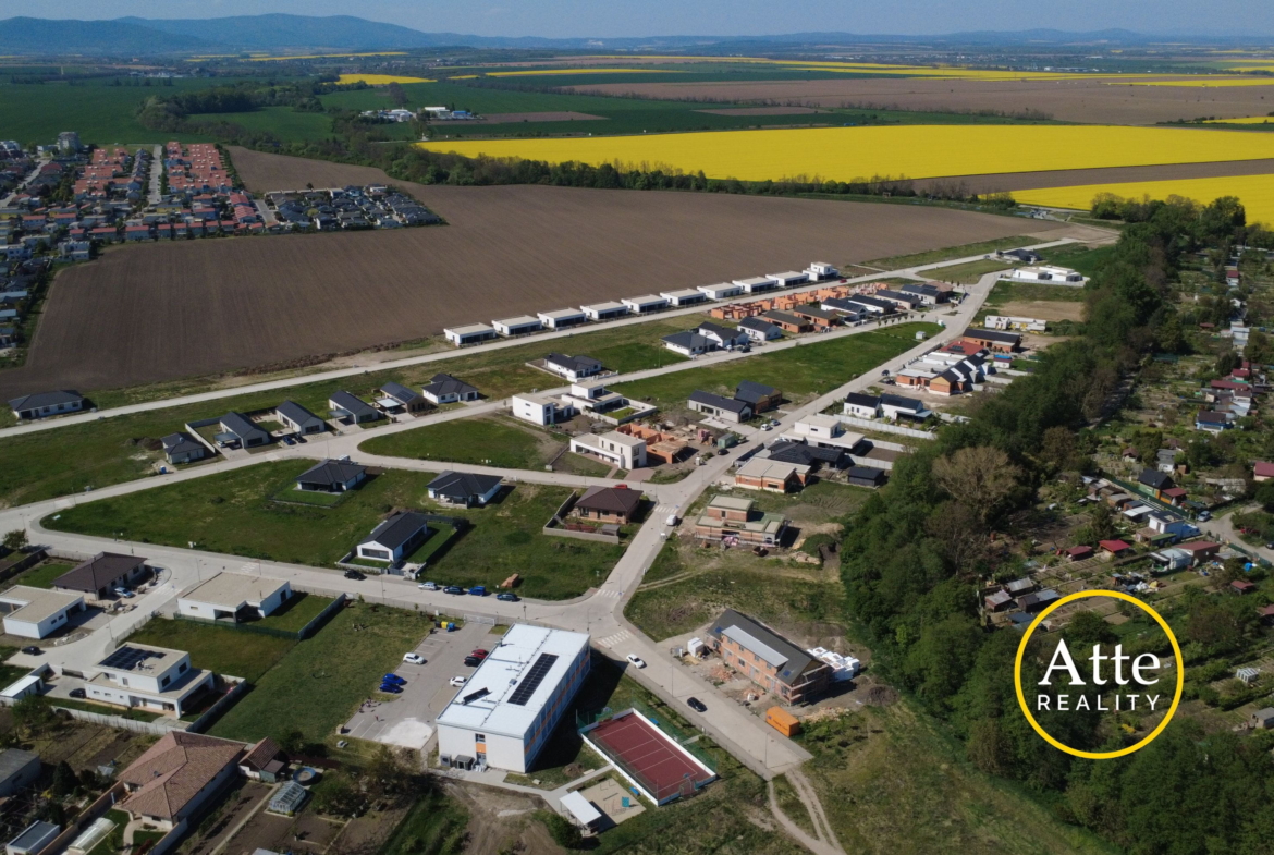 Aerial view of a developing suburb with rows of new houses along streets beside brown plowed fields and large yellow canola fields in the distance, with hills on the horizon. Real estate logo visible in the bottom-right corner.