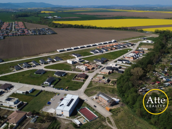 Aerial view of a developing suburb with rows of new houses along streets beside brown plowed fields and large yellow canola fields in the distance, with hills on the horizon. Real estate logo visible in the bottom-right corner.