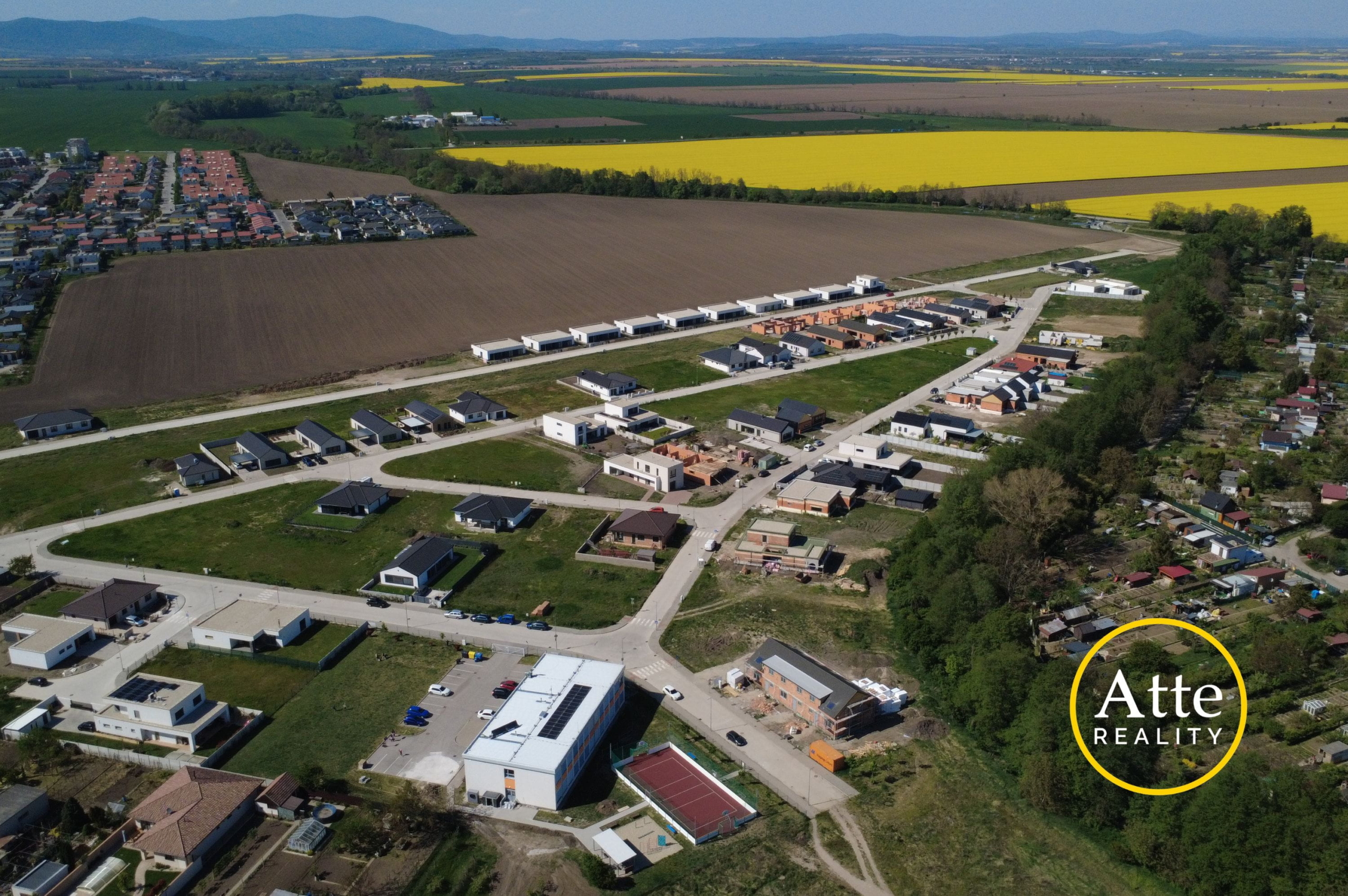 Aerial view of a developing suburb with rows of new houses along streets beside brown plowed fields and large yellow canola fields in the distance, with hills on the horizon. Real estate logo visible in the bottom-right corner.