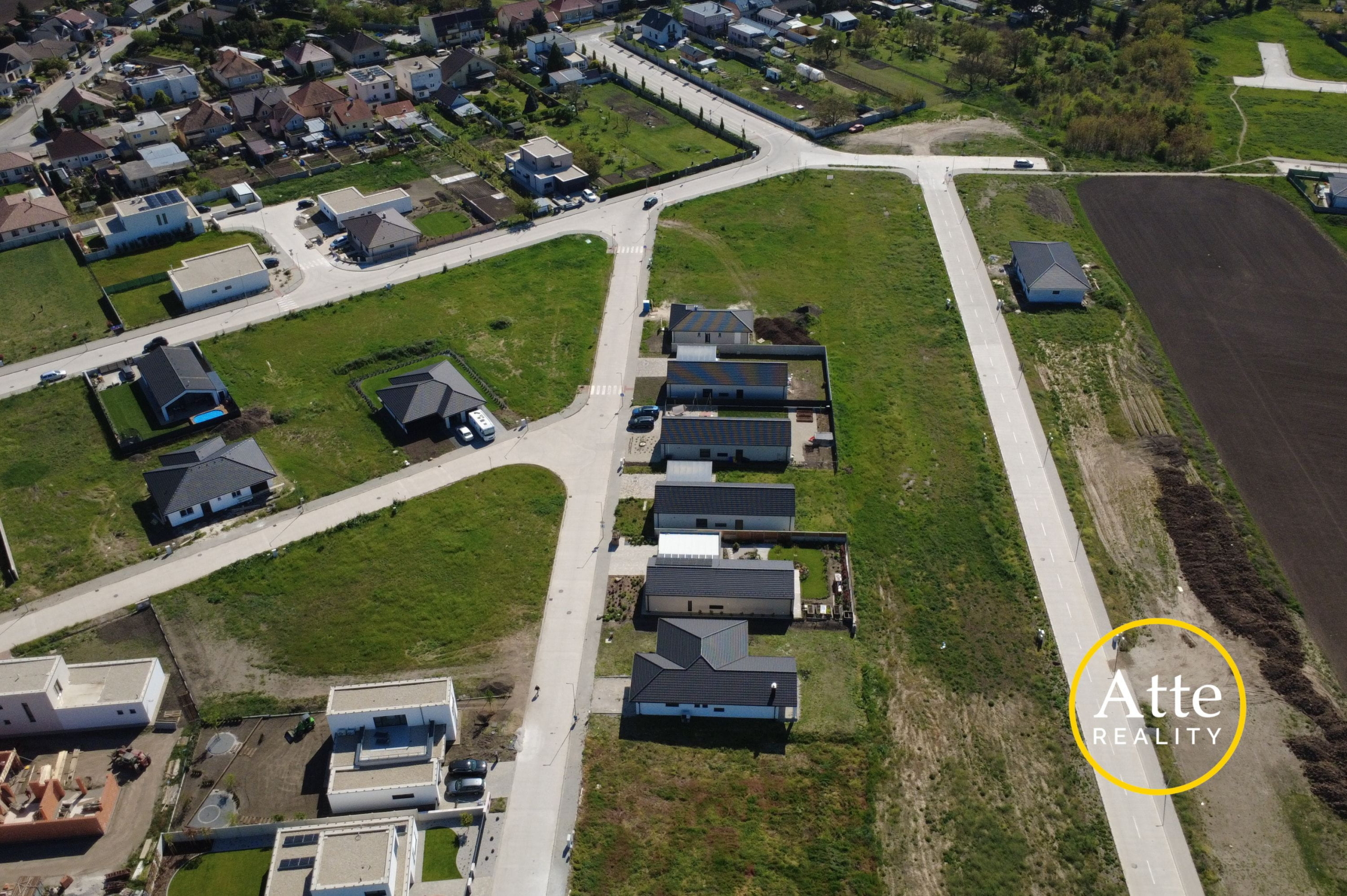 Aerial view of a suburban neighborhood with intersecting roads, rows of houses and gardens, and a large plowed field on the right; watermark reads Arte Realty.